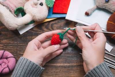 Hands crocheting a small strawberry, part of a creative and fun Crochet Kit project.